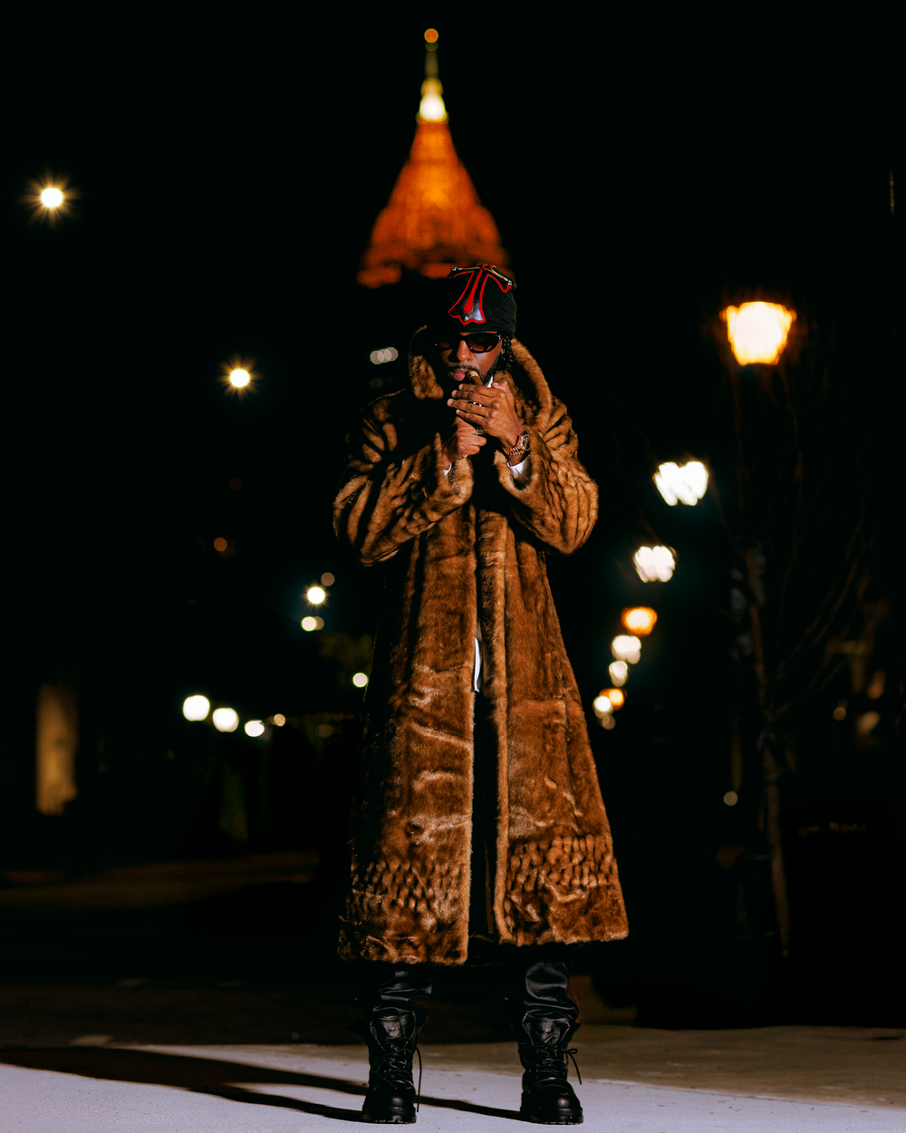 A performer in an ornate coat stands on stage at night with illuminated street lamps glowing in the background.