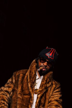 A person wearing a brown fur coat and black baseball cap poses against a dark background in a series of shots.