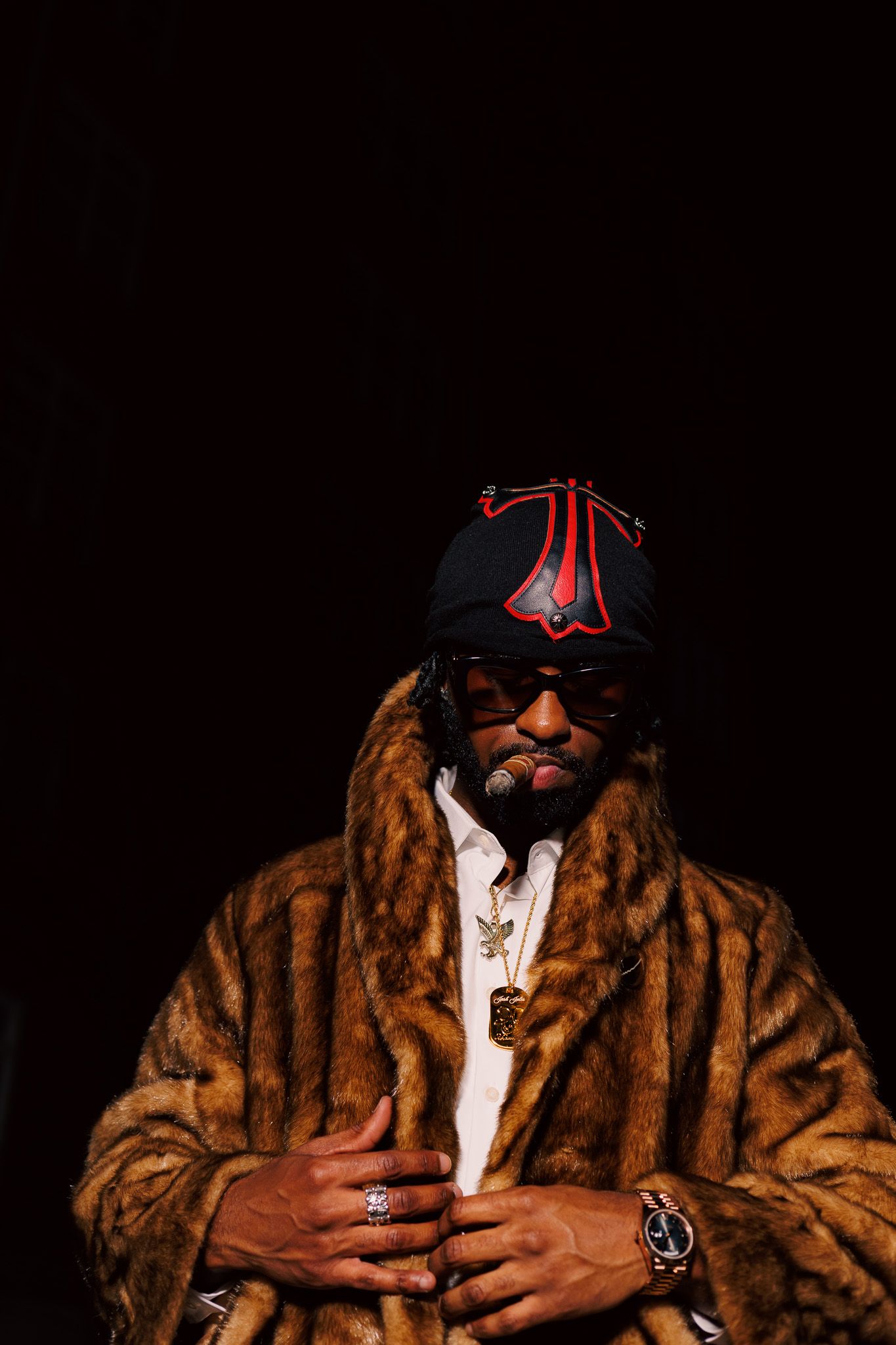 A person wearing sunglasses and a brown fur coat poses against a black background in a stylish fashion portrait series.
