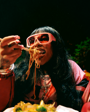 A person in sunglasses and pink clothing takes a bite of stringy melted cheese from a slice of pizza.