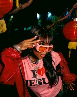 Nighttime fashion shot with pink sunglasses, red jacket and Jesus shirt amid colorful lantern decorations.