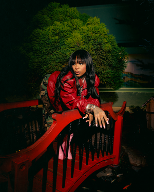 A dramatic fashion portrait in a dark garden setting with a figure in a striking red coat posed on wrought iron furniture.