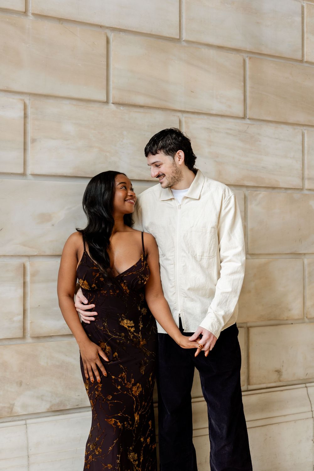 A couple shares a joyful moment together against a stone wall, with her wearing a floral dress and him in casual white.