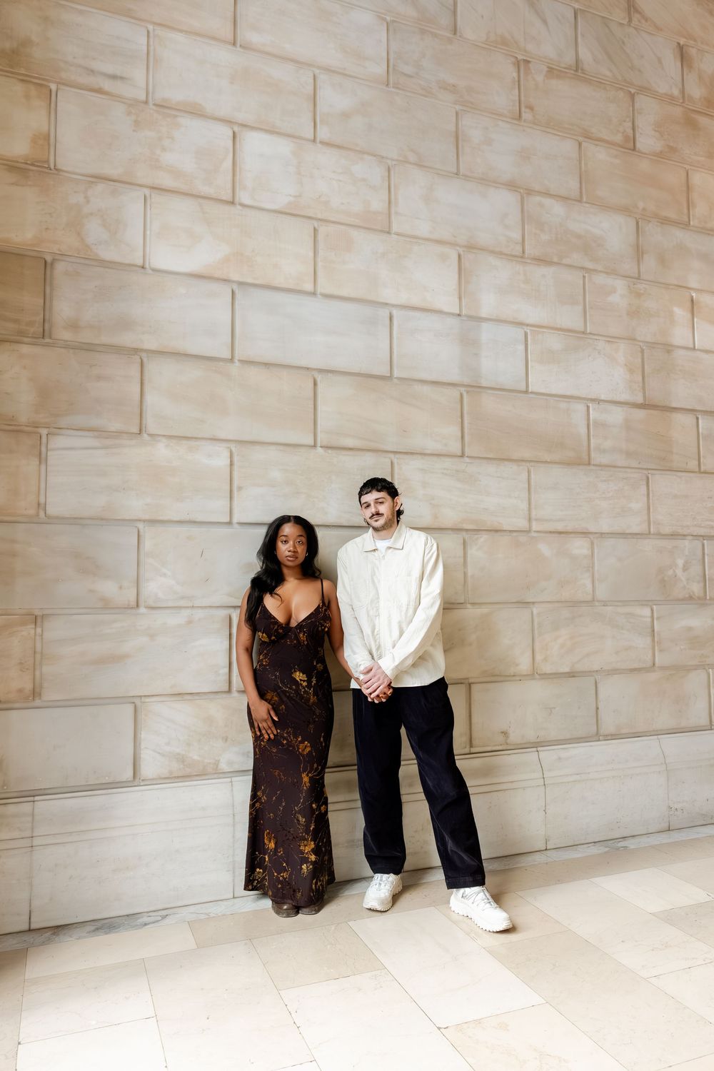 Two people pose against a beige stone wall in a creative fashion photography series.