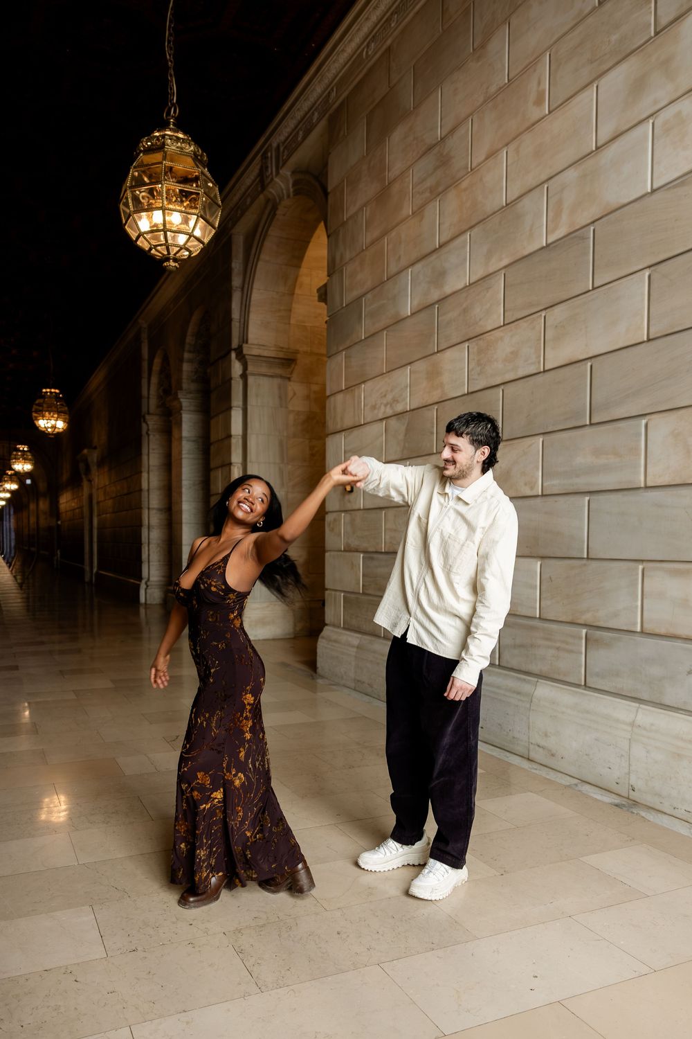 Two people dance together in an elegant hallway with stone walls and hanging lanterns creating a romantic atmosphere.