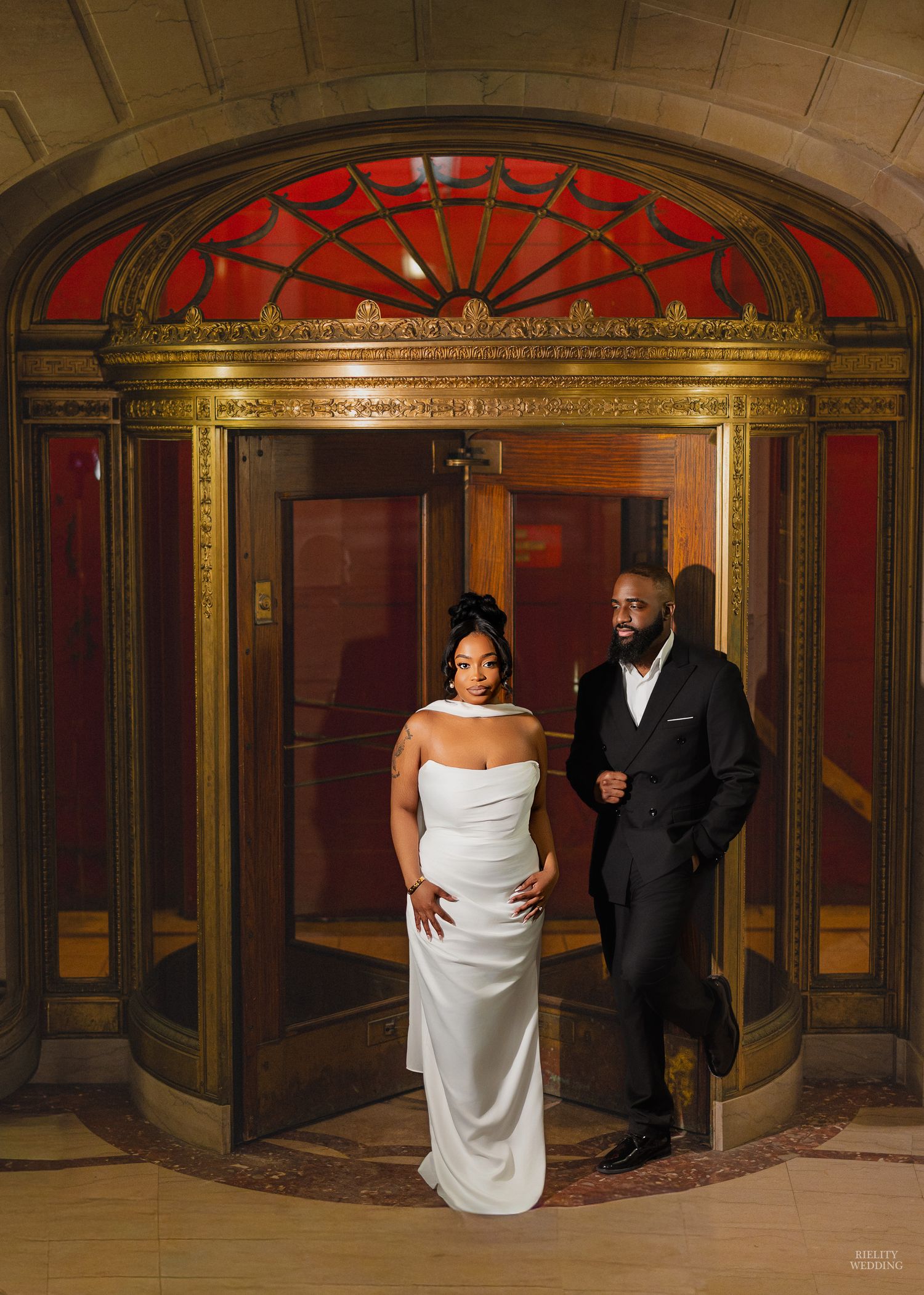 Elegant wedding couple poses in ornate vintage hallway with red and gold architectural details and arched windows.