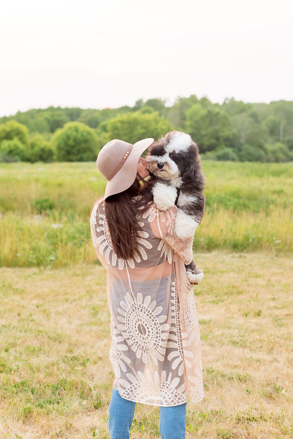 A black and white dog plays with someone wearing a beige hat and lace top in a sunny meadow.