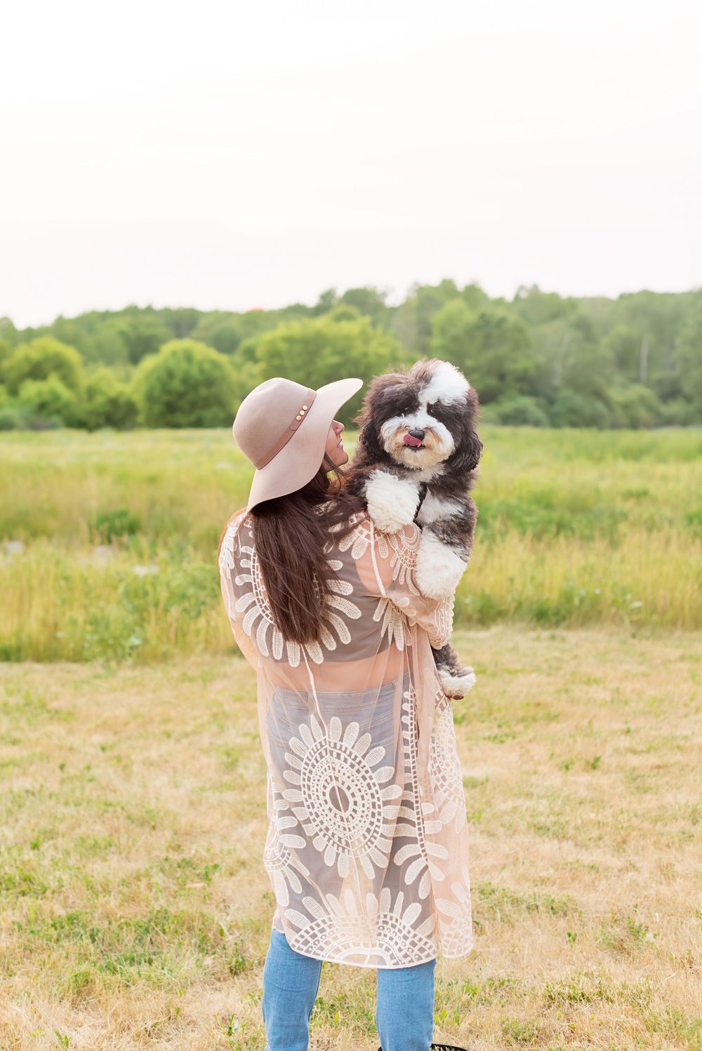 A black and white dog plays with someone wearing a beige hat and lace top in a sunny meadow.