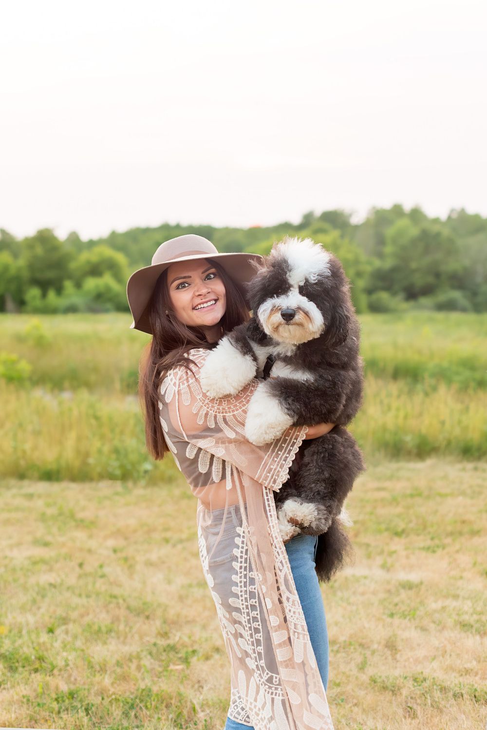 A black and white Bernedoodle dog being held outdoors by someone wearing a brown hat and lace dress.