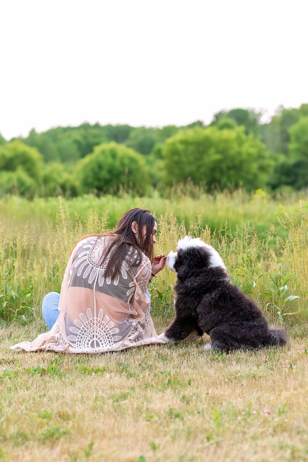 Person in lace top lying in grass field with black and white dog on summer day.