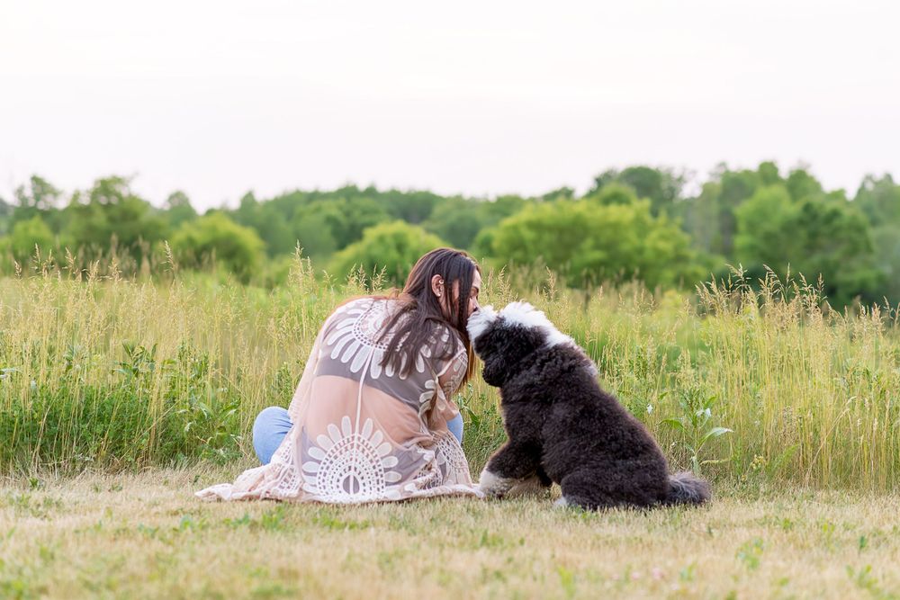 A person and black dog lying together in a grassy field on a sunny summer day.