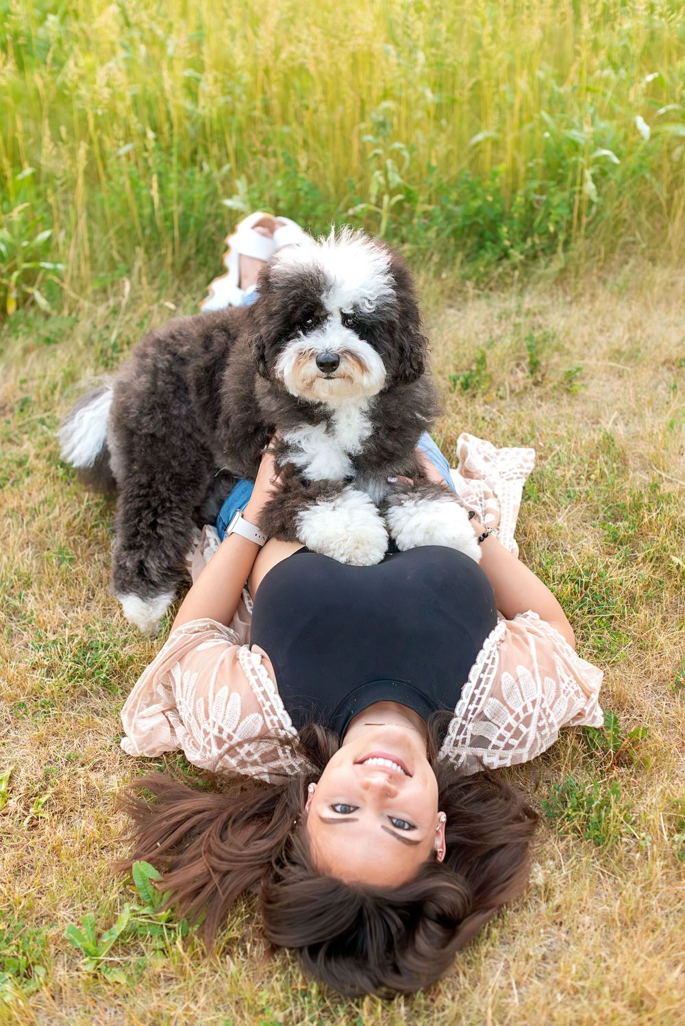 Person in lace dress lying on grass with fluffy black and white dog enjoying sunny day.