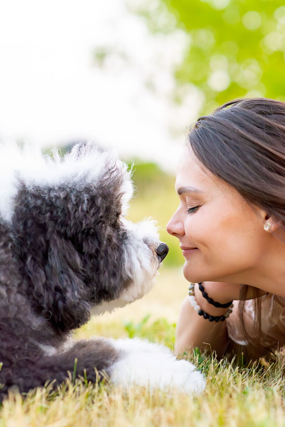A person and a fluffy black and white dog share a tender moment lying in grass with soft bokeh background.