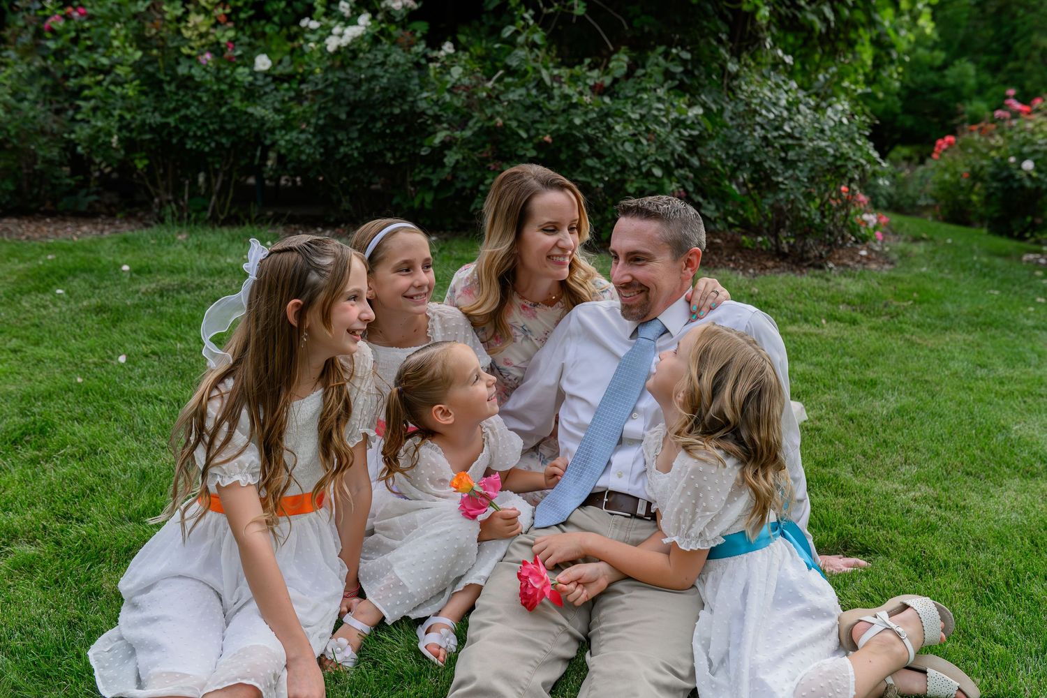 A family of 5 sits together in the College of Southern Idaho rose garden for candid family photos. They direct natural smiles towards dad.