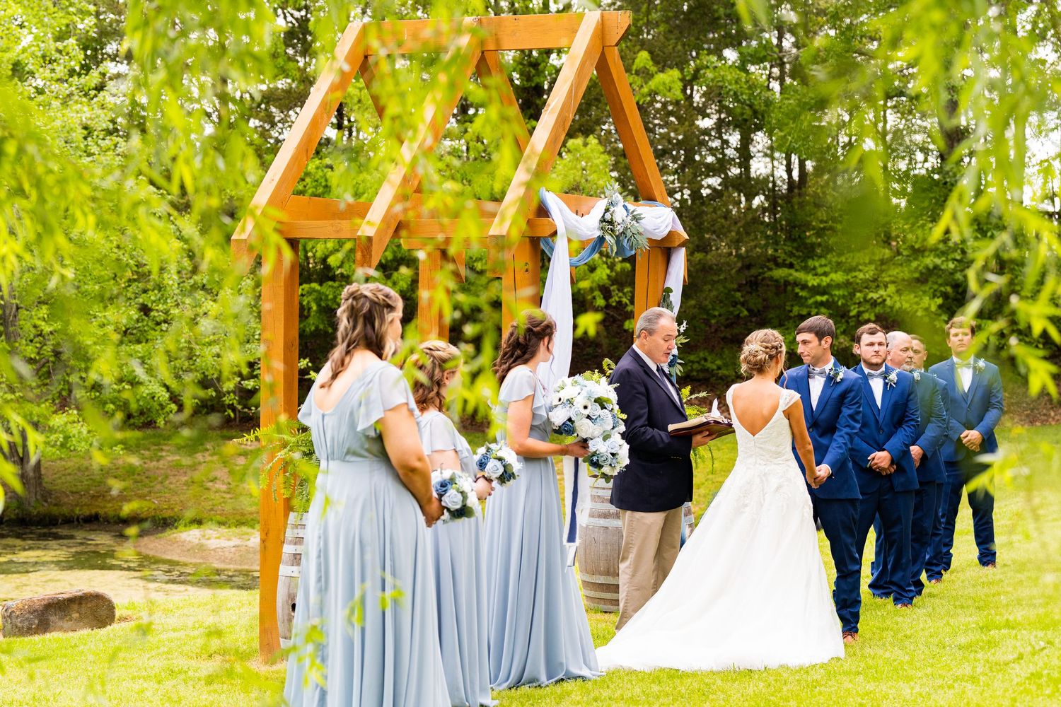 Wedding party in light blue bridesmaid dresses and navy suits poses together in a lush green outdoor garden setting.