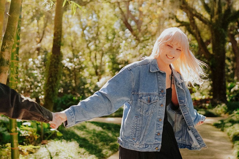 Person in denim jacket walks along tree-lined path during autumn season with golden sunlight filtering through leaves.