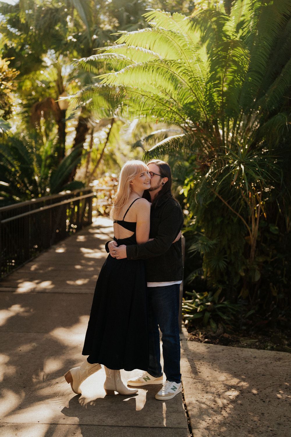 A couple embraces on a garden pathway surrounded by lush ferns and tropical foliage in golden sunlight.