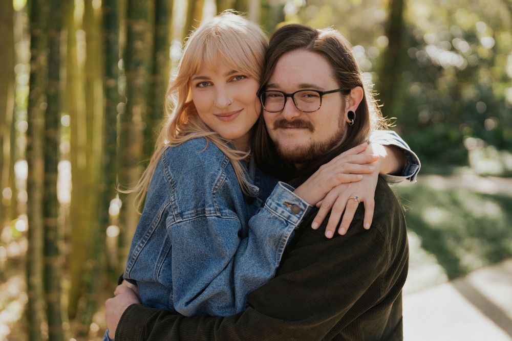 A couple embraces and smiles together in a sunlit forest setting wearing casual denim and dark clothing.