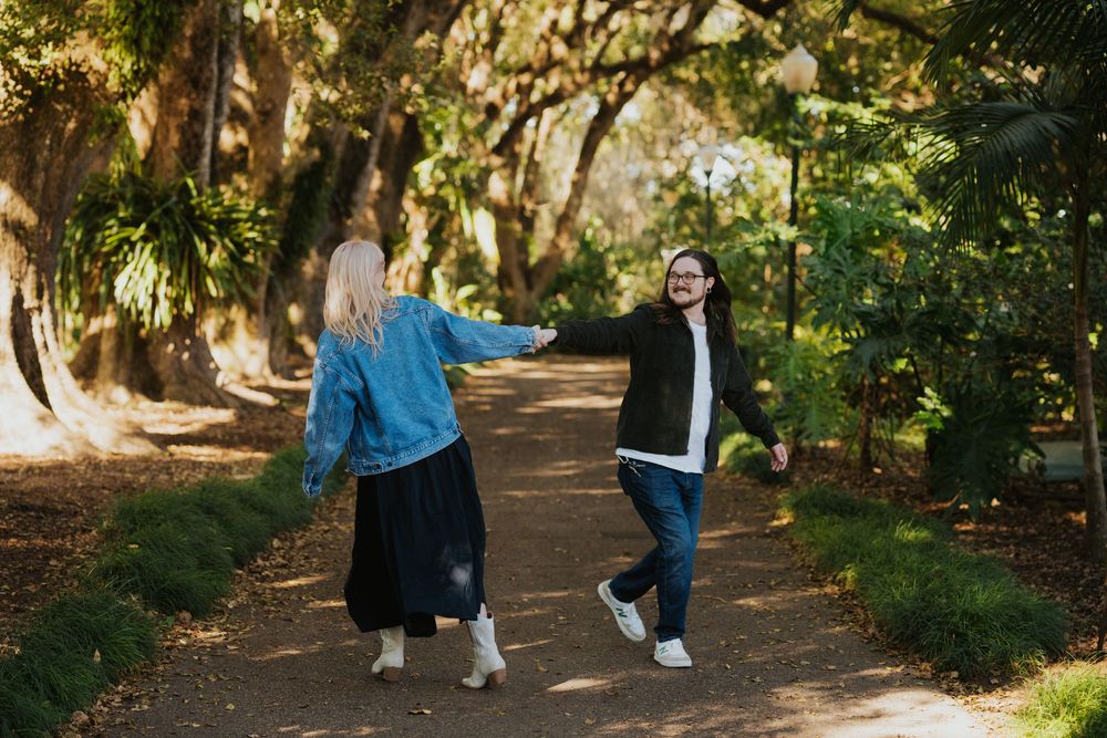 Two people walking together and laughing on a tree-lined path in a park on a sunny day.