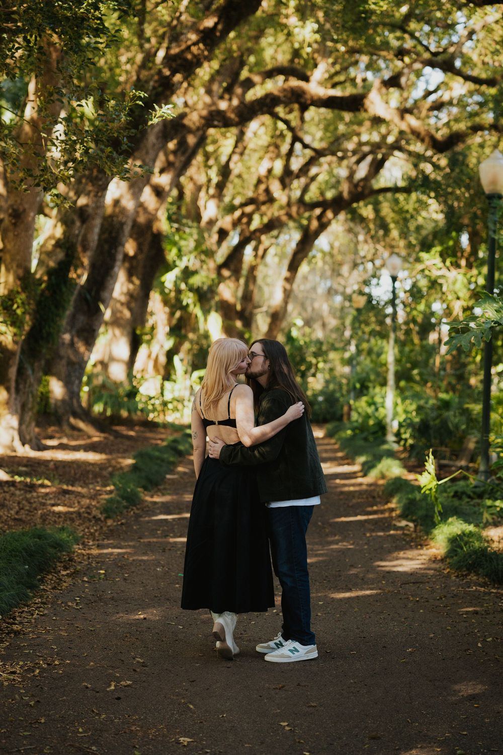 A romantic couple shares intimate moments along a tree-lined dirt path in a sunlit forest setting.