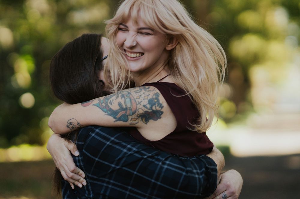 Two people share a joyful embrace outdoors with visible tattoo art and blonde hair against a sunlit natural background.