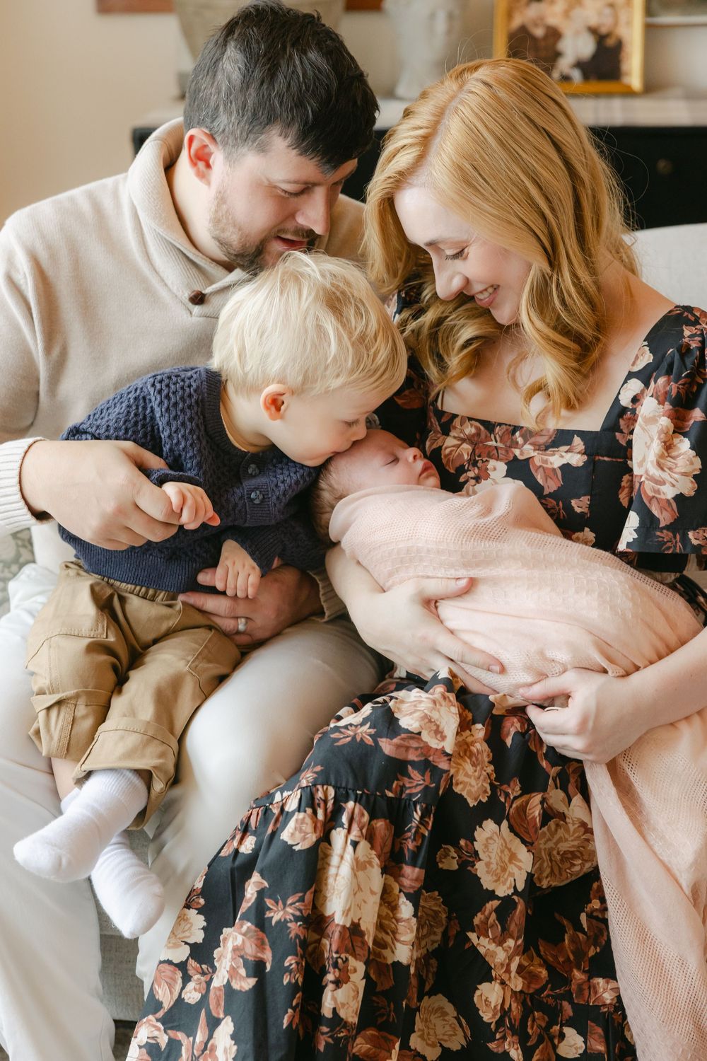 A family sits together on a couch, with a toddler in khaki pants leaning in to look at their newborn sibling.  In-home Newborn Session CT