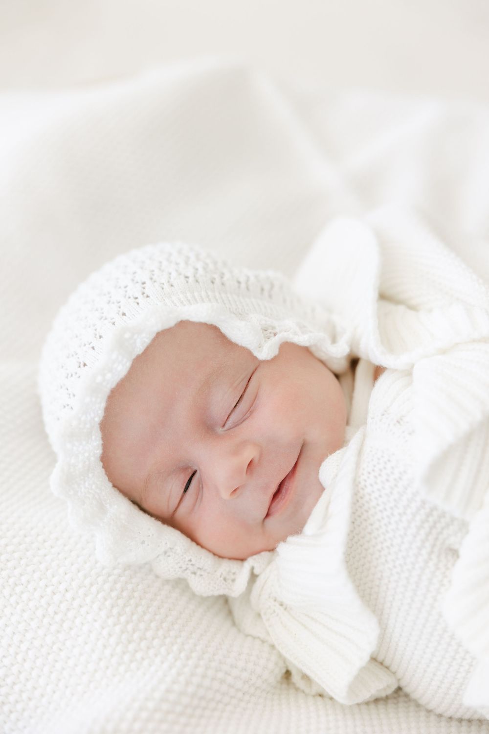 A sleeping baby wearing a white bonnet and wrap lies peacefully on textured white bedding. Studio Newborn Photographer