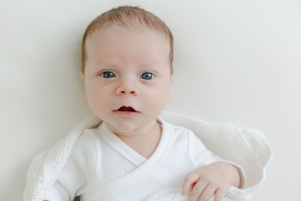 A baby wearing a white onesie lies on a soft white blanket while looking at the camera with bright eyes. Studio CT Newborn Photographer