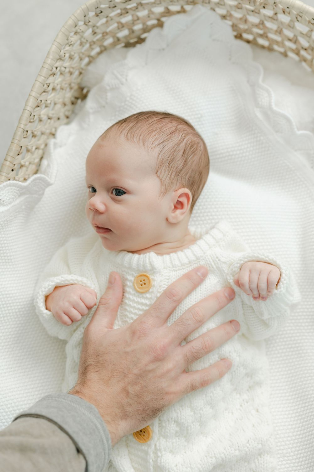 A baby wearing a white knit sweater lies on a delicate lace blanket while an adult's hand provides gentle support. Studio Newborn Photograph