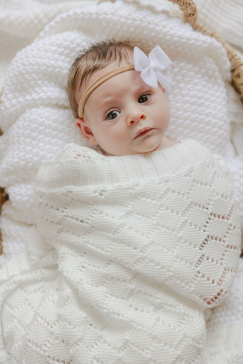 Baby wearing white bow headband lies wrapped in soft knitted white blanket with delicate diamond pattern. In-home Newborn Session CT