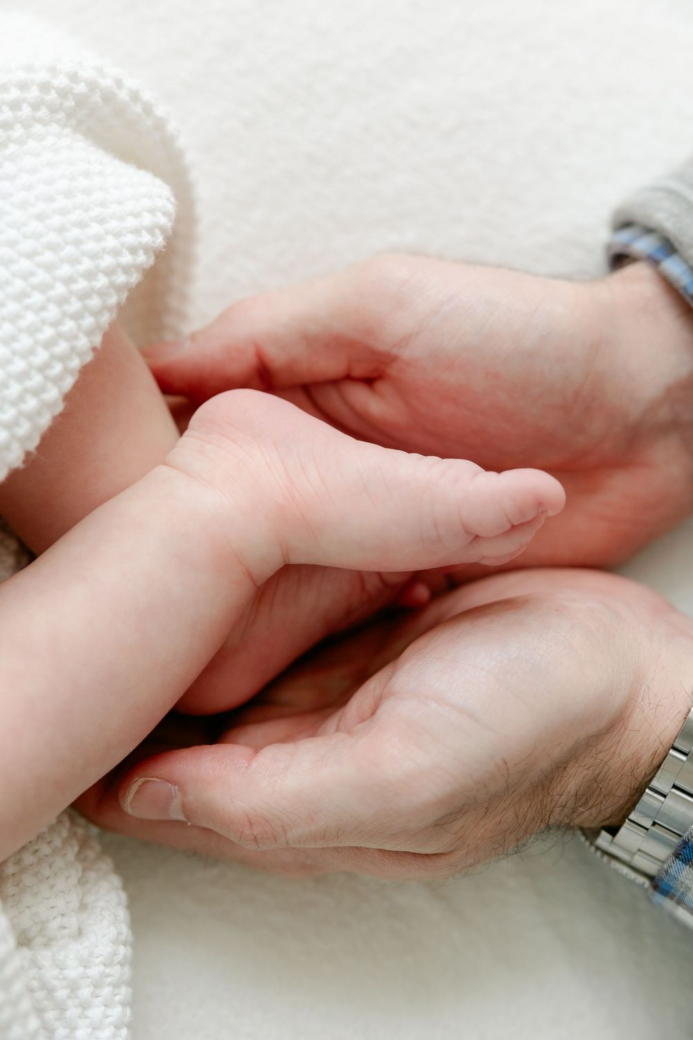 Two hands gently clasped together against a white textured background, with one person wearing a silver watch. Studio Newborn Photographer