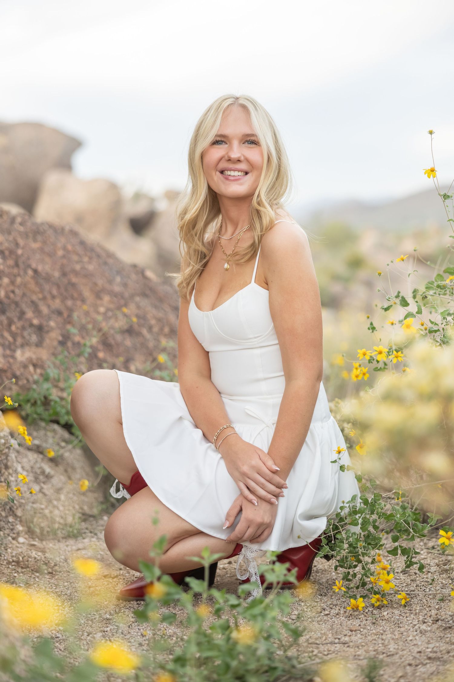 A person in a white summer dress sits among yellow wildflowers in a desert landscape on a sunny day.