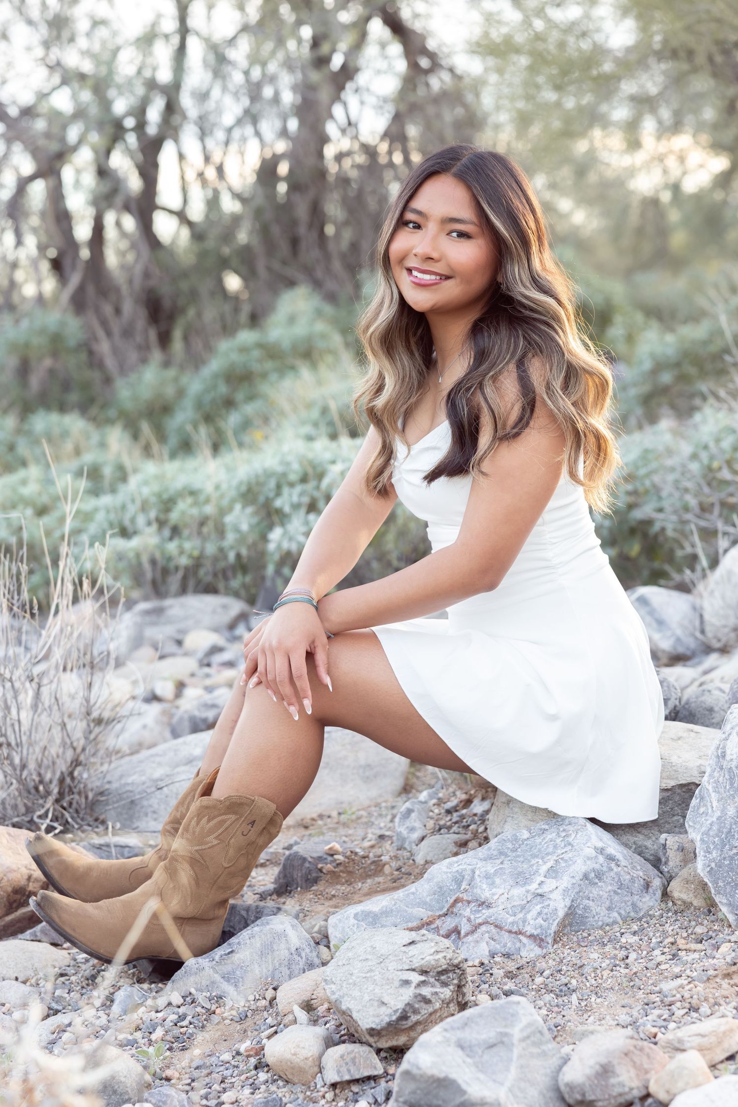 Woman in white dress and brown cowboy boots poses on rocky terrain in a desert landscape with soft natural lighting.