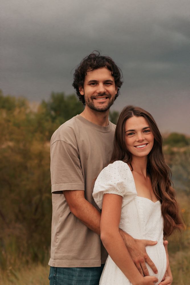 Smiling couple outdoors, man in tan shirt embracing pregnant woman in white dress | Westminster Colorado Family Photographer