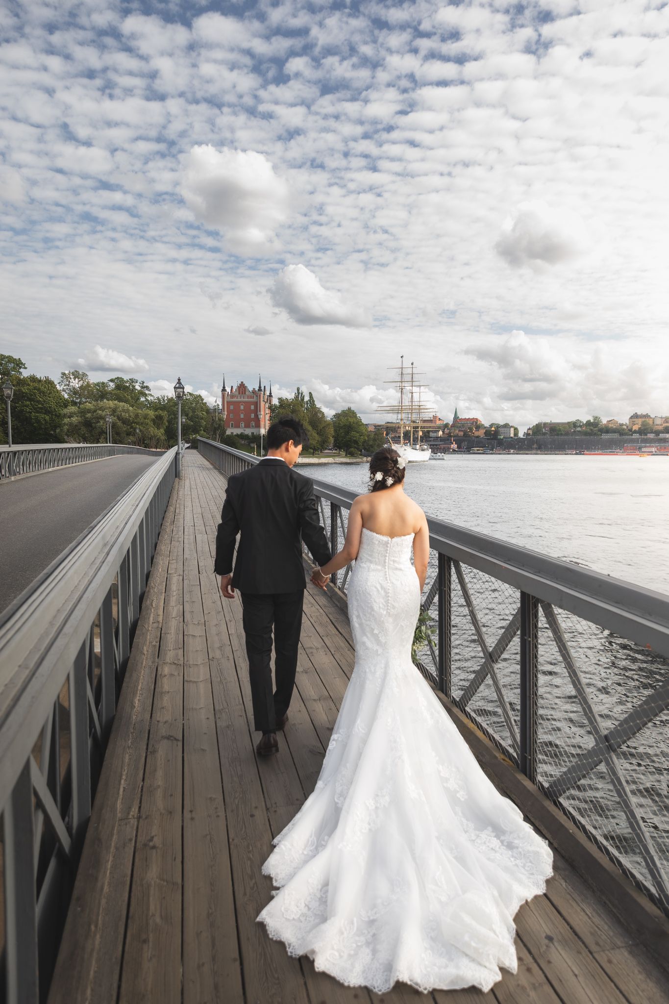 A couple in formal wedding attire walks along a wooden pier with a cloudy sky and waterfront backdrop.