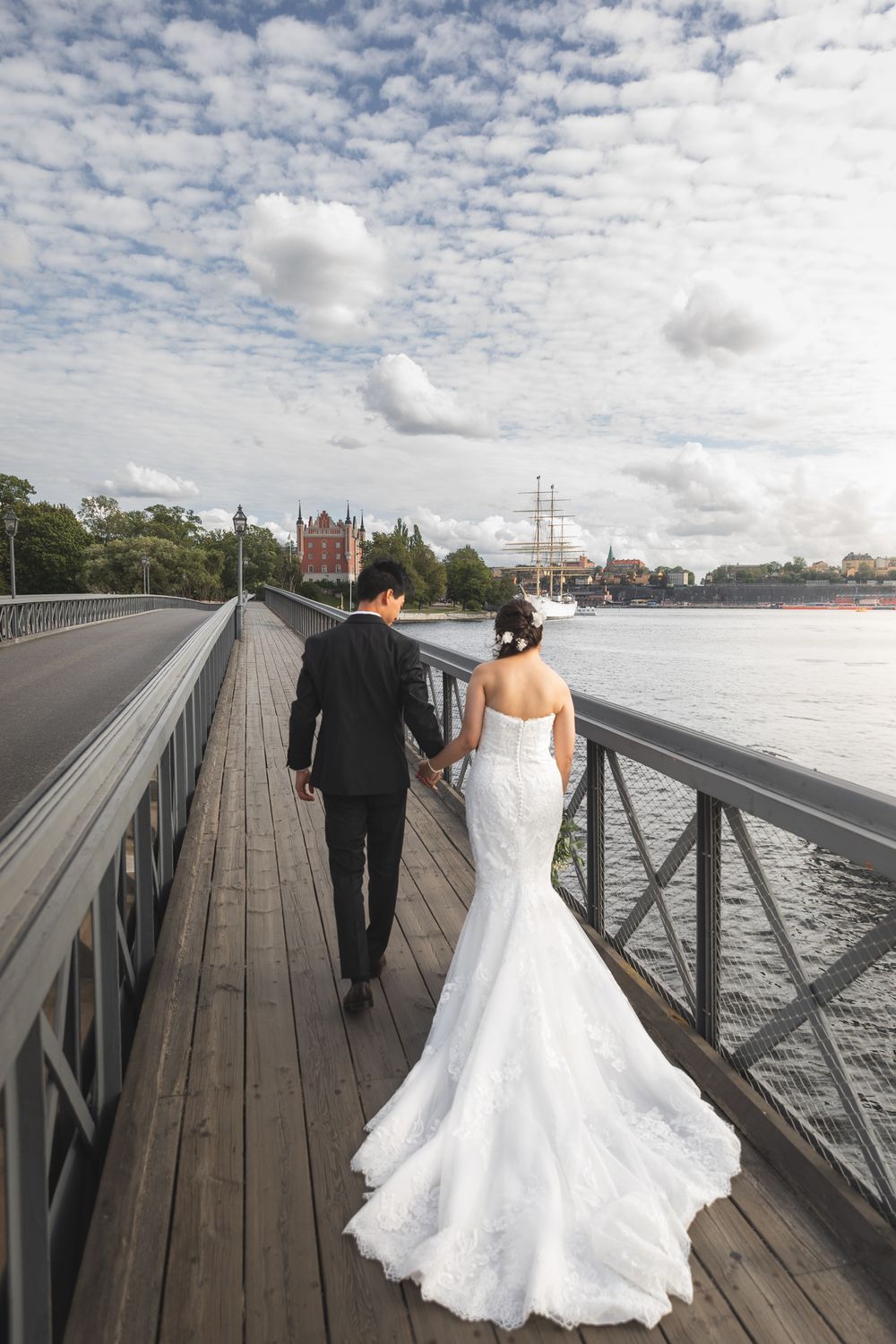 A couple in formal wedding attire walks along a wooden pier with a cloudy sky and waterfront backdrop.