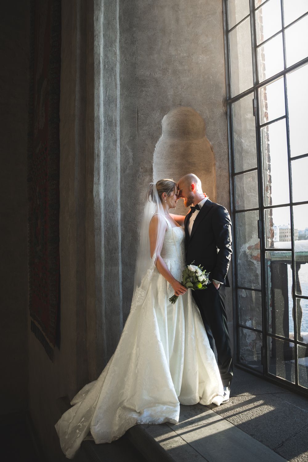 A romantic wedding couple poses by large windows in an elegant venue with natural light streaming in.