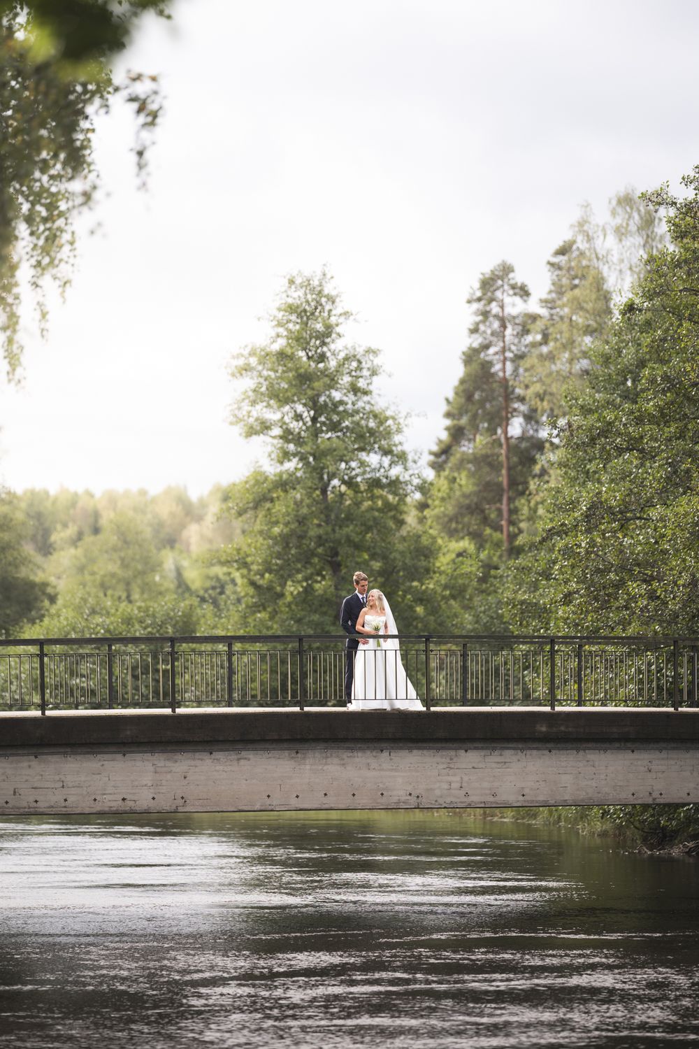A couple shares a romantic moment on a wooden bridge over rippling water surrounded by lush green trees.