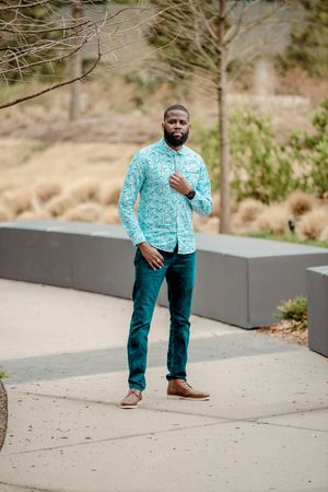 Person wearing turquoise patterned shirt and teal pants posing outdoors in casual setting.