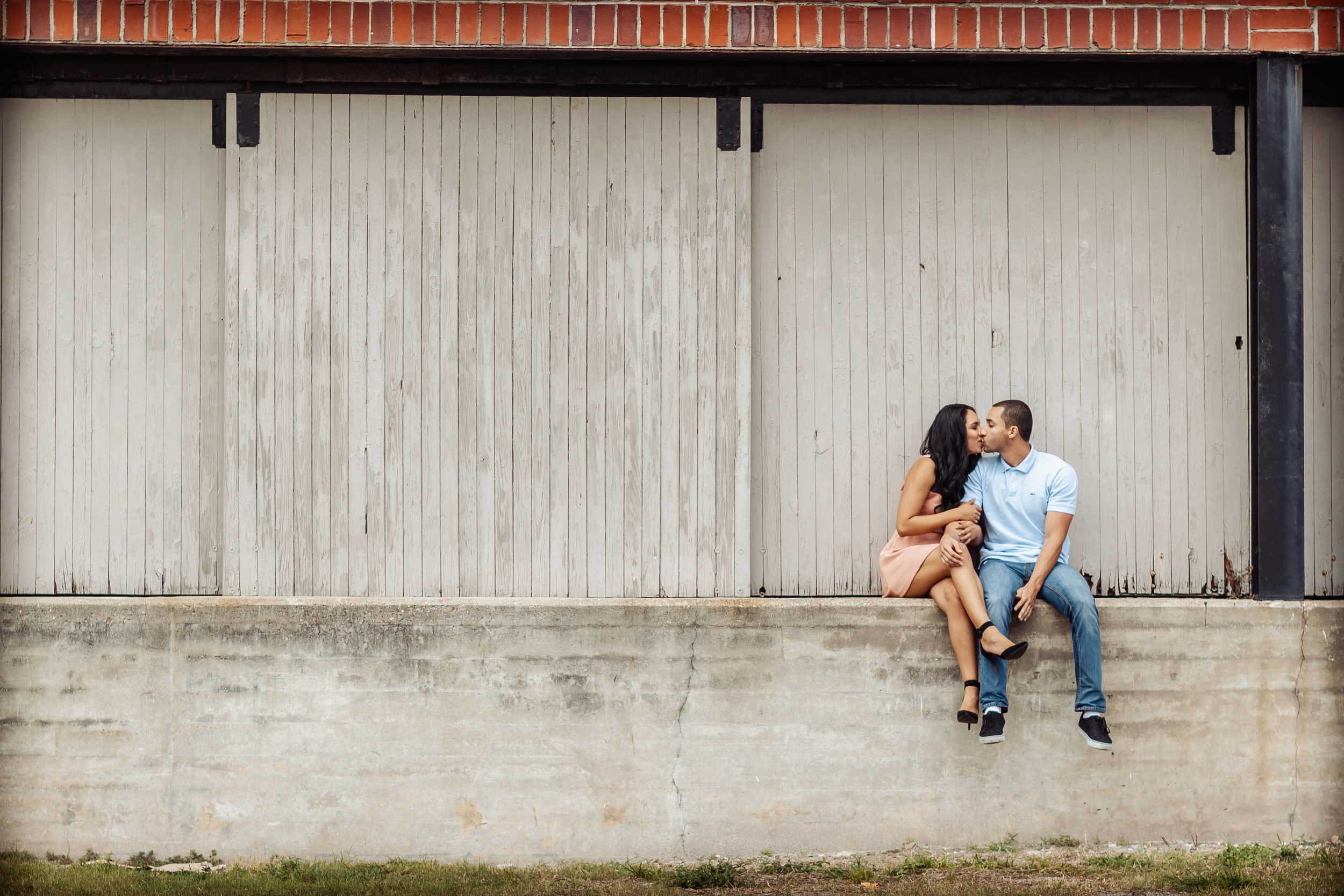 Couple poses against industrial wall in romantic engagement photo series.