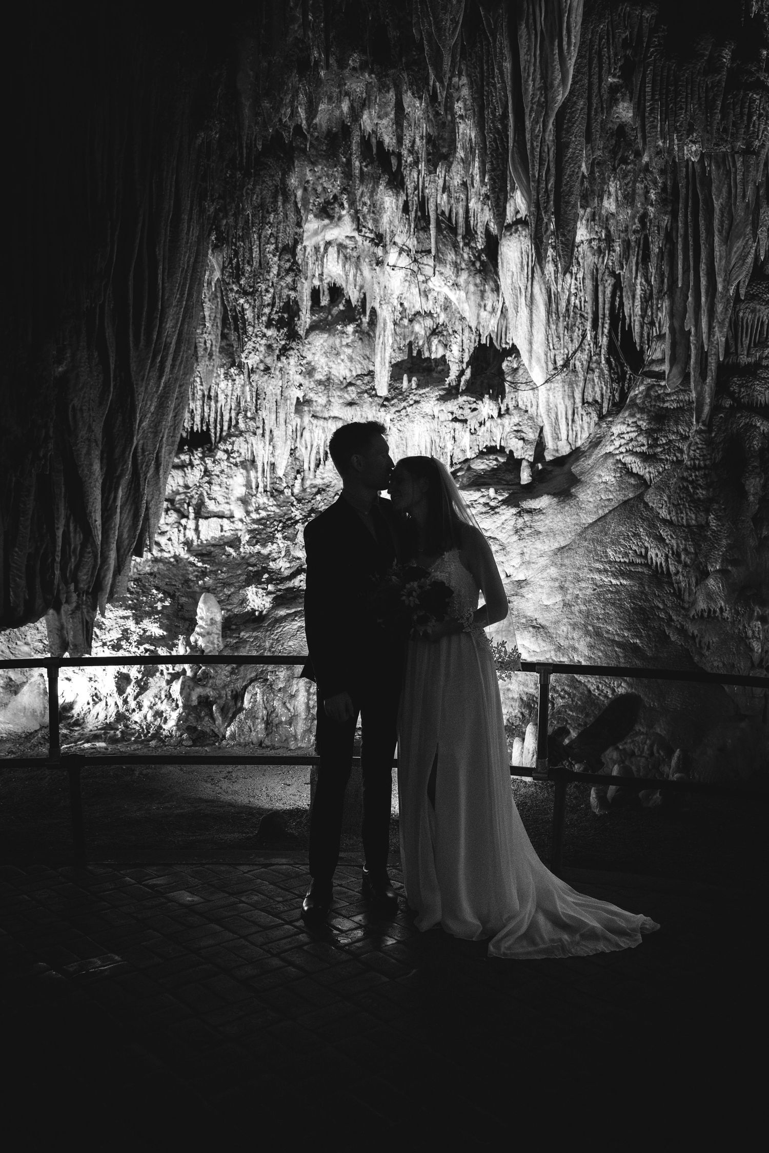 A romantic couple poses in silhouette against dramatic cave formations lit by spotlights in an underground cavern.