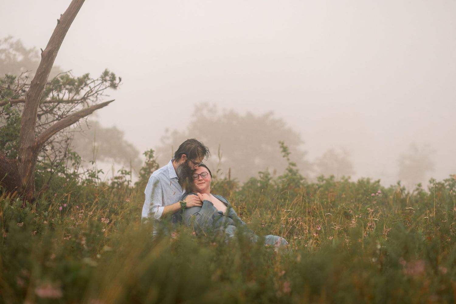 Couple sitting in a misty field surrounded by lush greenery, sharing a tender romantic moment in foggy outdoor setting.