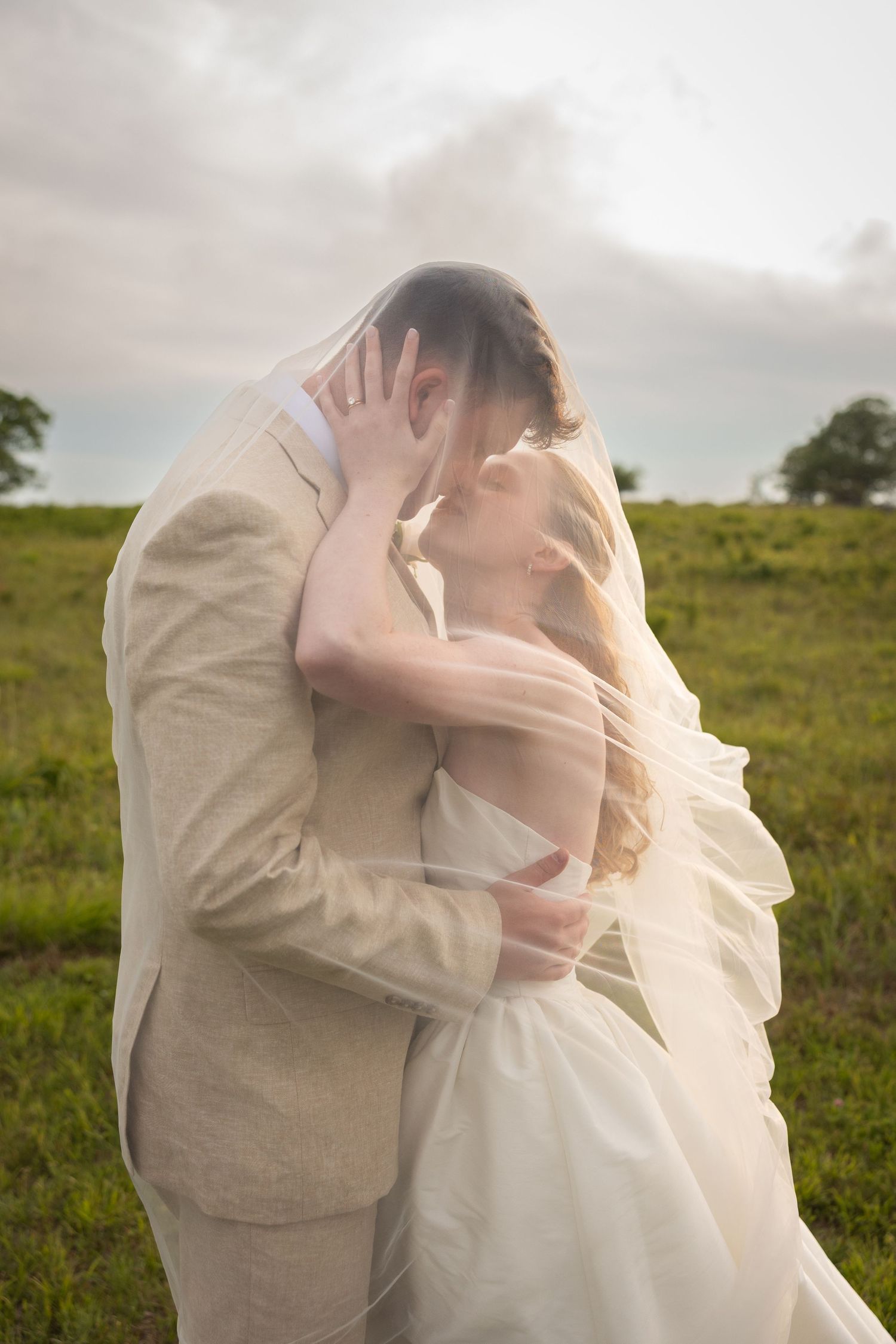 Bride and groom sharing a romantic kiss under a veil in a lush green field on their wedding day.