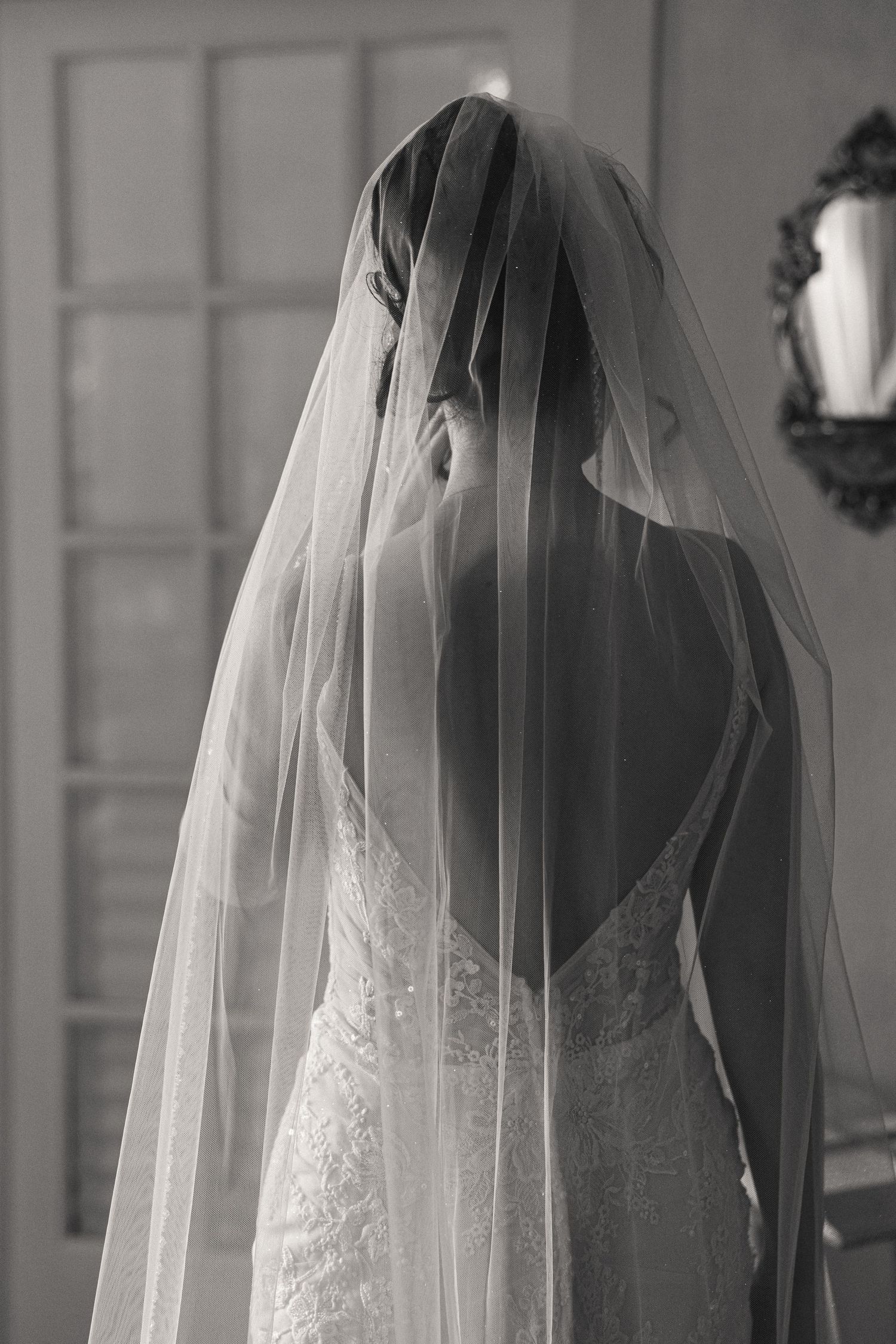 Black and white photo of a bride in a lace wedding dress with a flowing veil, viewed from behind.