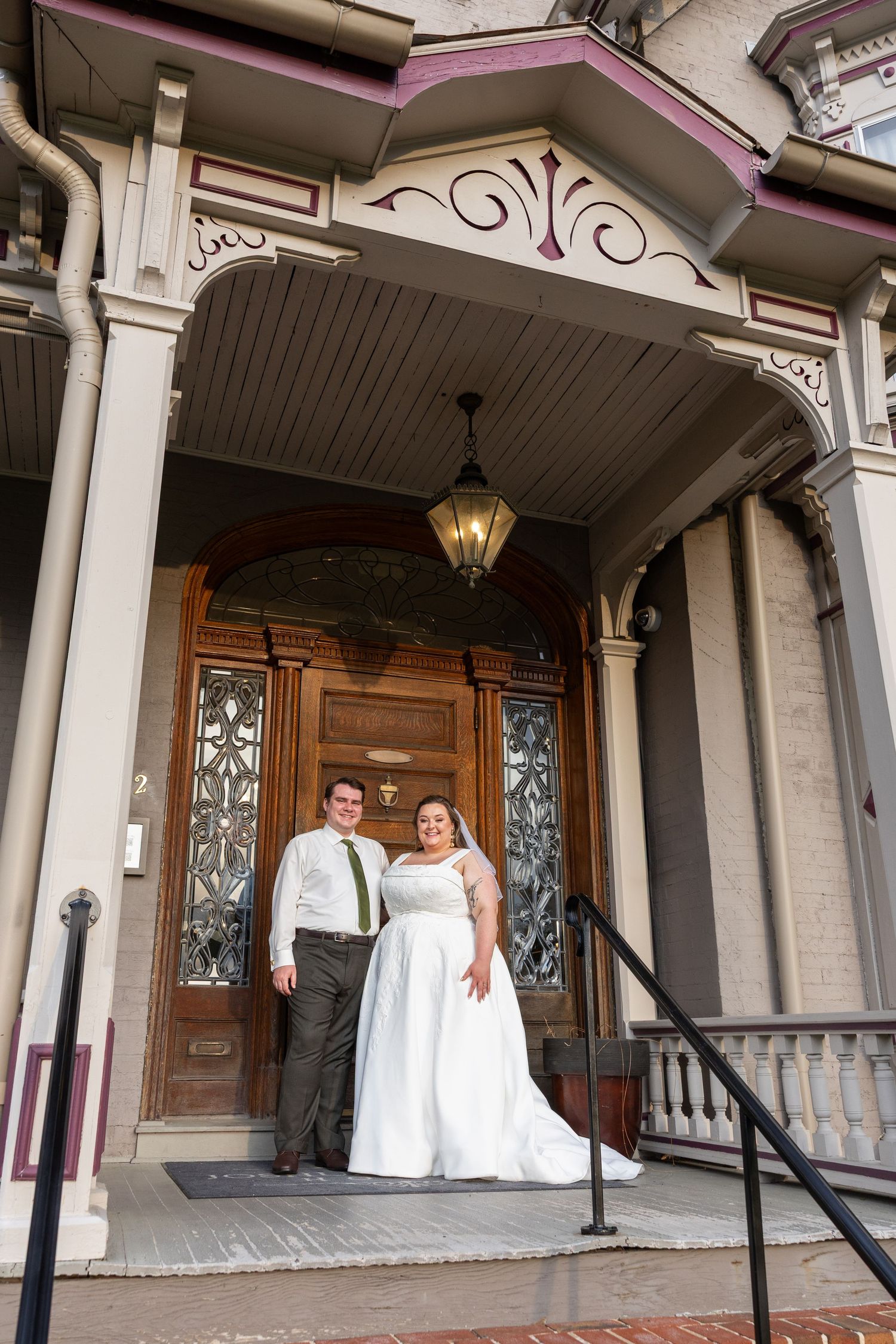 Bride and groom posing on ornate Victorian building entrance steps, bride in white gown, groom in white shirt with green tie.