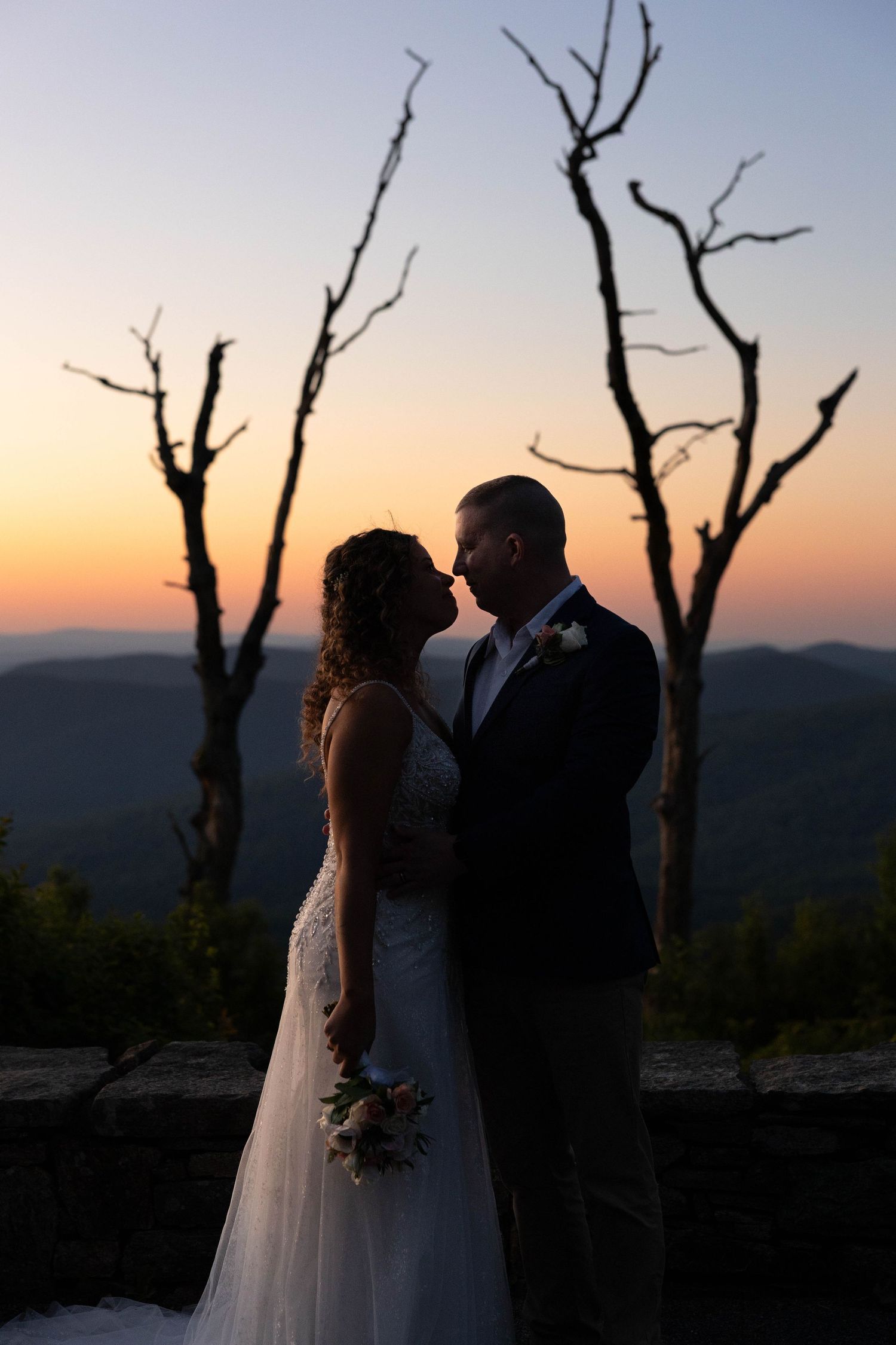 Bride and groom silhouetted against a stunning sunset with bare trees and mountain views at their romantic outdoor wedding.
