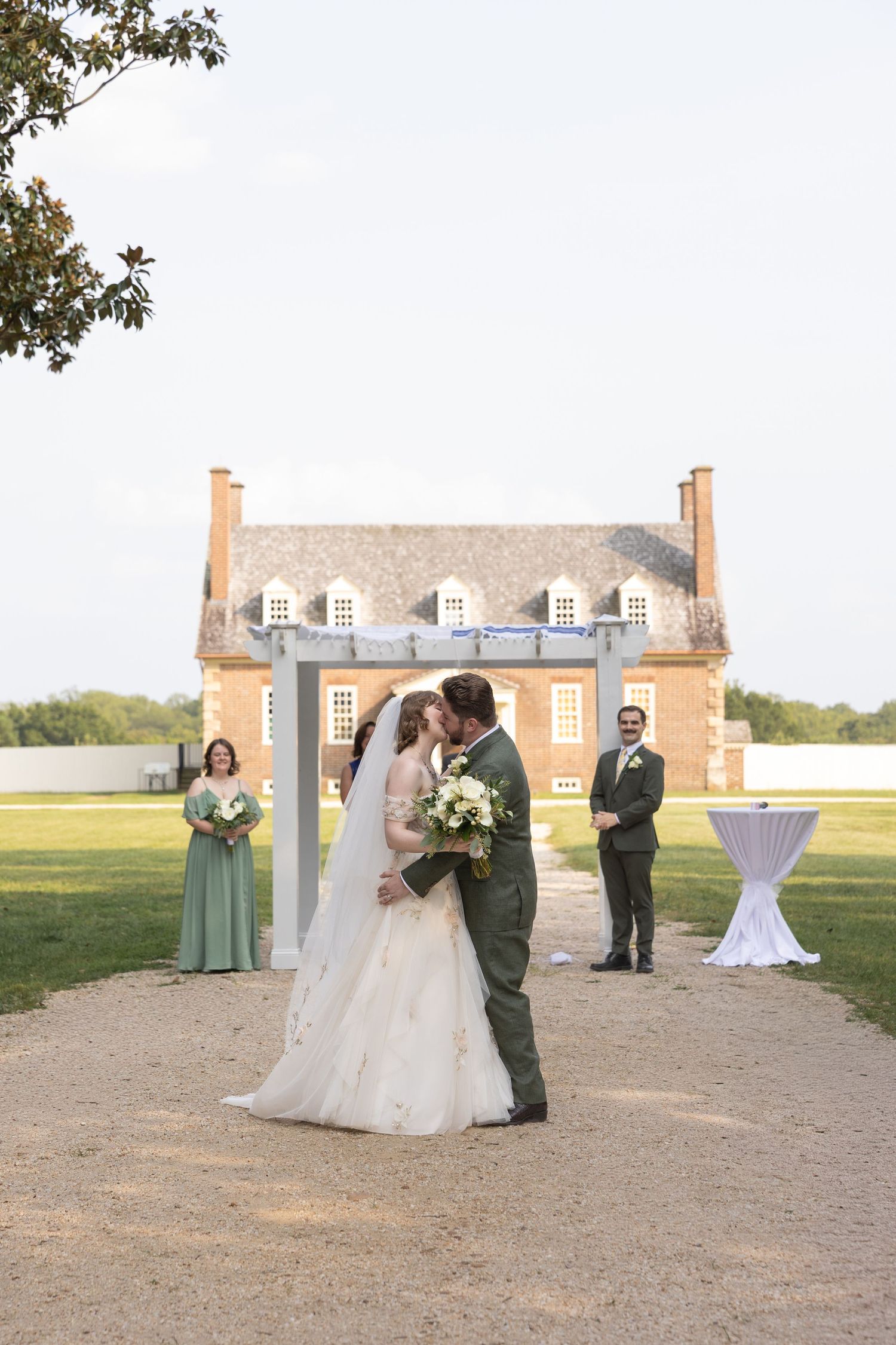 Bride and groom share first kiss at outdoor wedding ceremony in front of historic brick building with wedding party.