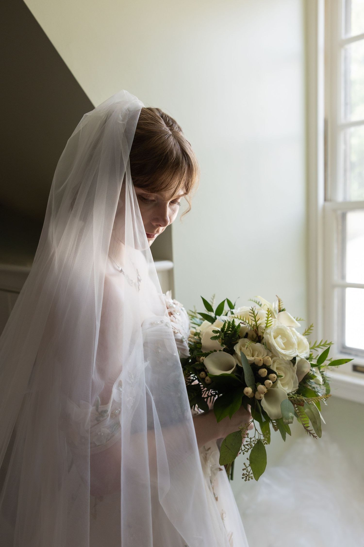 Bride in white lace gown and veil holds elegant bouquet of white roses and greenery near a sunlit window.
