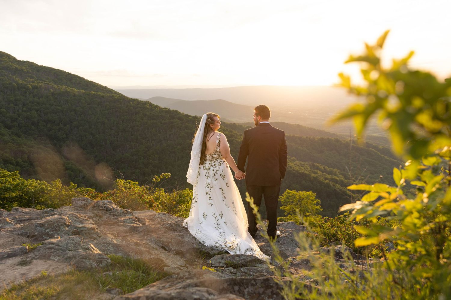 Bride and groom holding hands on rocky mountain overlook at sunset with lush green valley views.