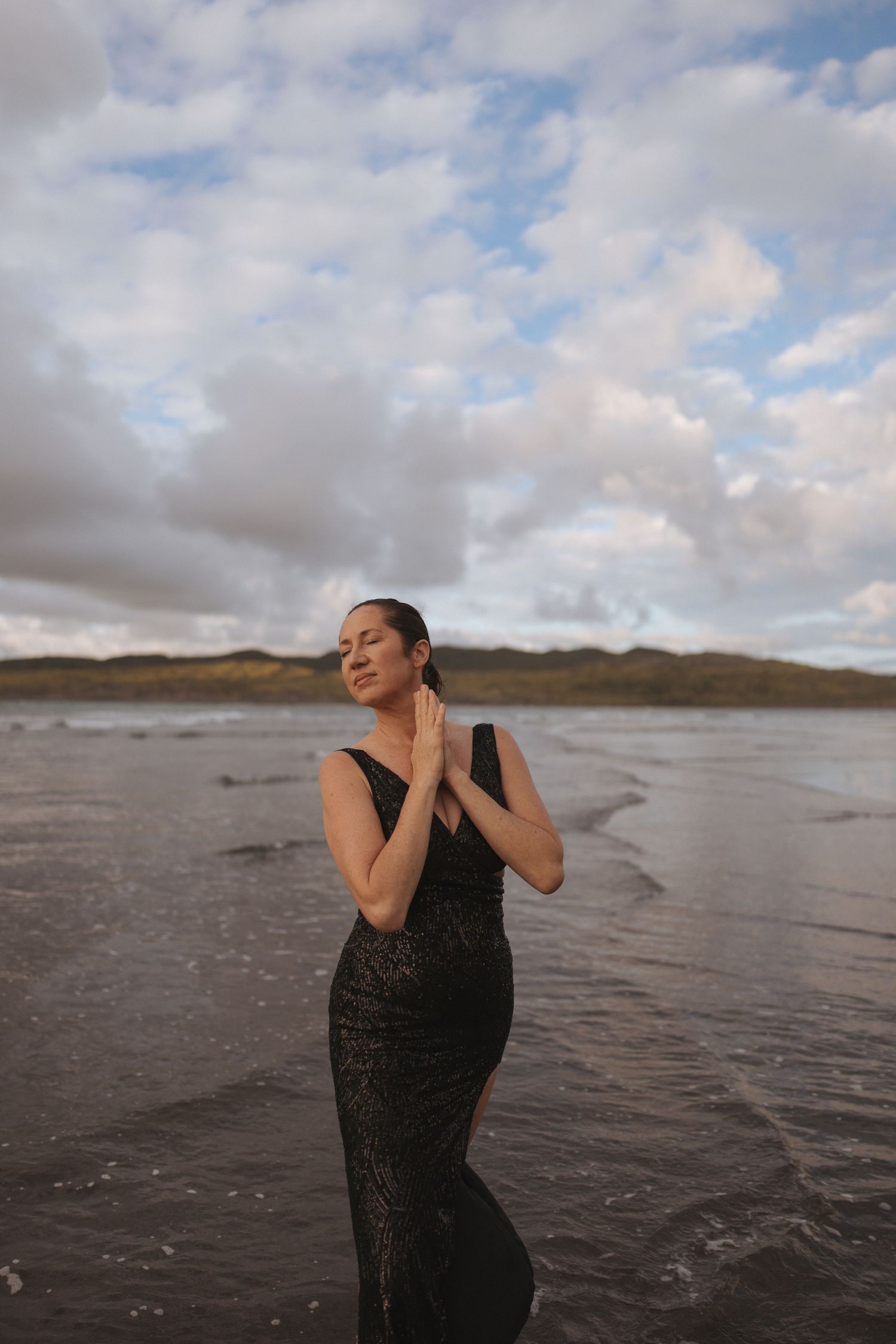 Woman in black lace gown standing in ocean waves with eyes closed under dramatic cloudy sky in New Zealand.